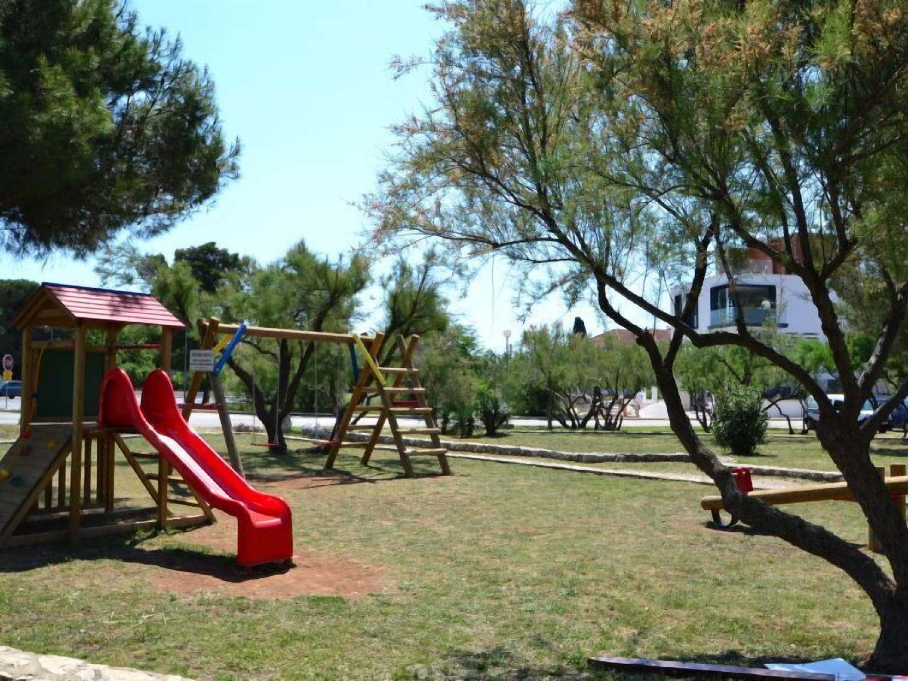 Un parque infantil con un tobogán rojo y columpios, rodeado de árboles y un edificio al fondo.