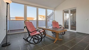 A balcony with two rocking chairs and a table, large windows, and a tiled floor.