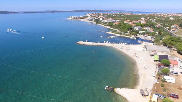 An aerial view of a coastal town with clear blue waters, a sandy beach, and a marina with boats.