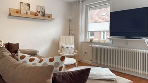 Ein modernes Wohnzimmer mit einer Couch, einem Sessel, einem Fernseher, einer Heizung und einem Fenster mit Blick auf ein Backsteingebäude.