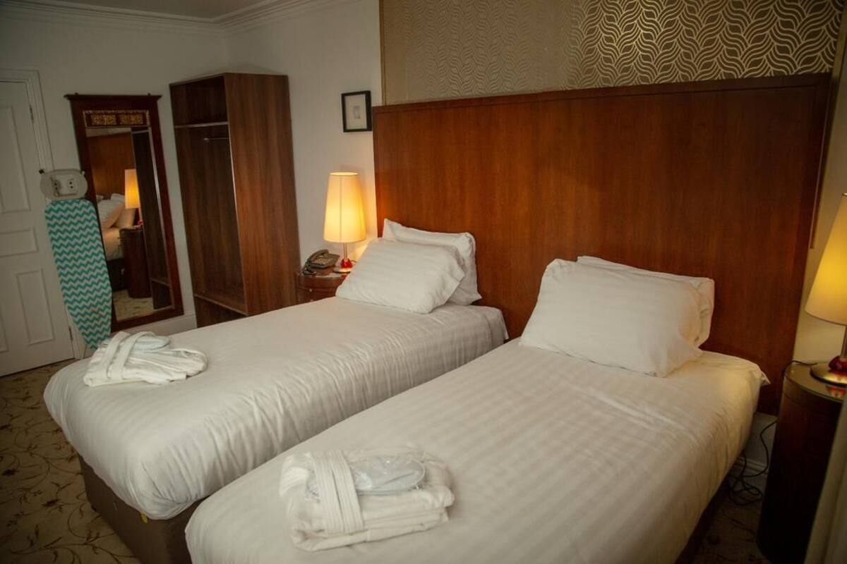 Hotellihuone, jossa on kaksi erillistä sänkyä, puinen pääty, yöpöytä lampun kanssa ja peili.