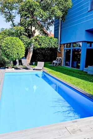 A swimming pool with a wooden deck, surrounded by a well-maintained lawn and a large tree. - Řeporyje by Interhome (Reporyje)