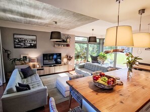 A modern living room with a sofa, television, and a dining area with a wooden table and fruit bowl.