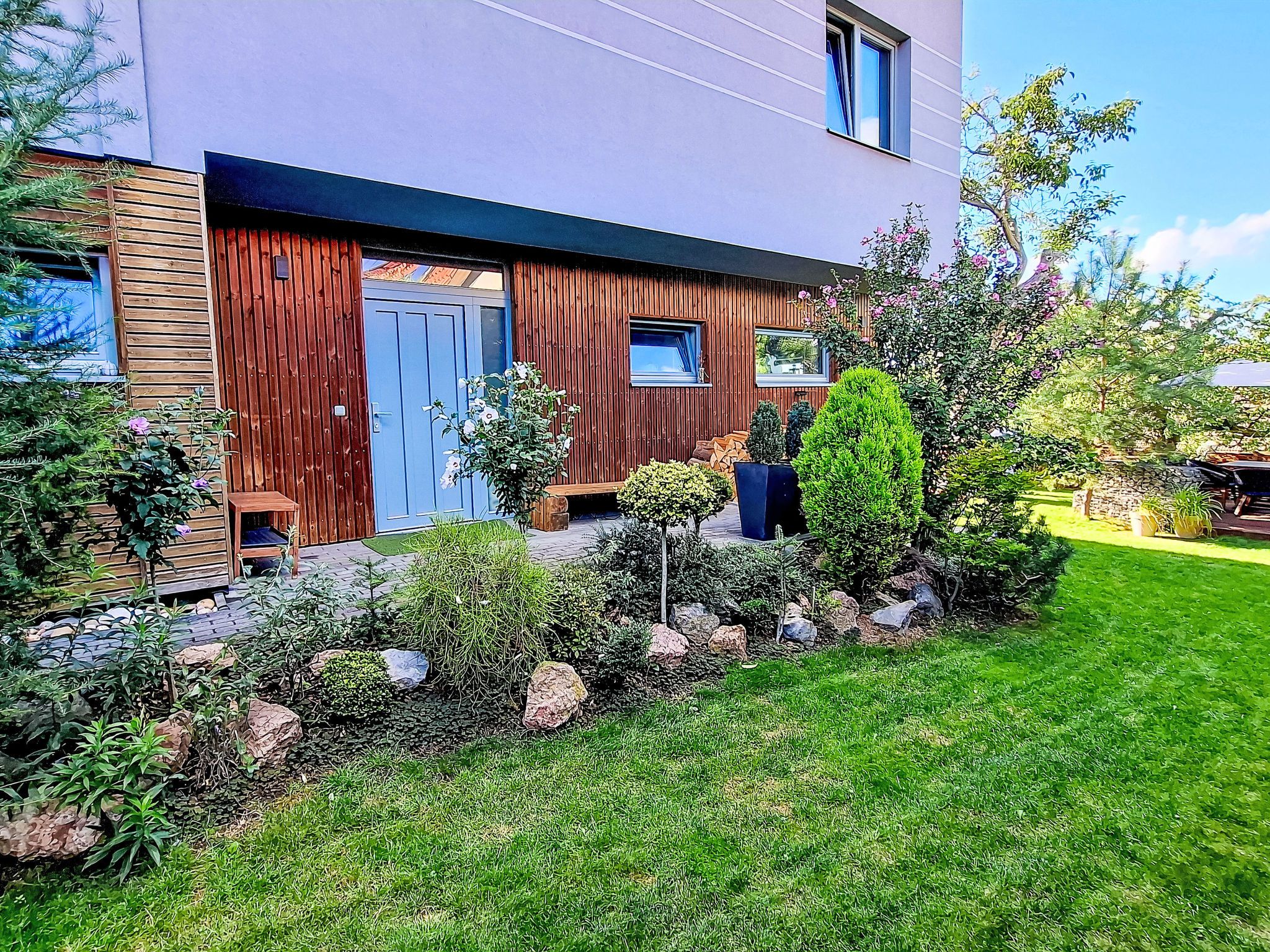 A modern house with a wooden facade, a blue door, and a well-maintained garden with various plants and a lawn.