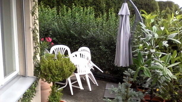 Ein kleiner Patio-Bereich mit einem weißen Stuhl und einem Tisch, umgeben von Topfpflanzen und einem weißen Sonnenschirm.