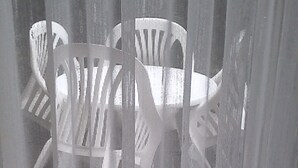 Blick durch transparente Vorhänge auf einen weißen Sitzbereich mit Stühlen und einem Tisch.