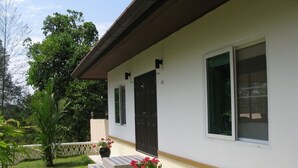 Ein Haus mit weißer Fassade, einer dunkelbraunen Tür und einem Fenster mit weißem Rahmen. Vor dem Haus befindet sich ein kleiner Garten mit einem Blumentopf und einem gepflasterten Weg.