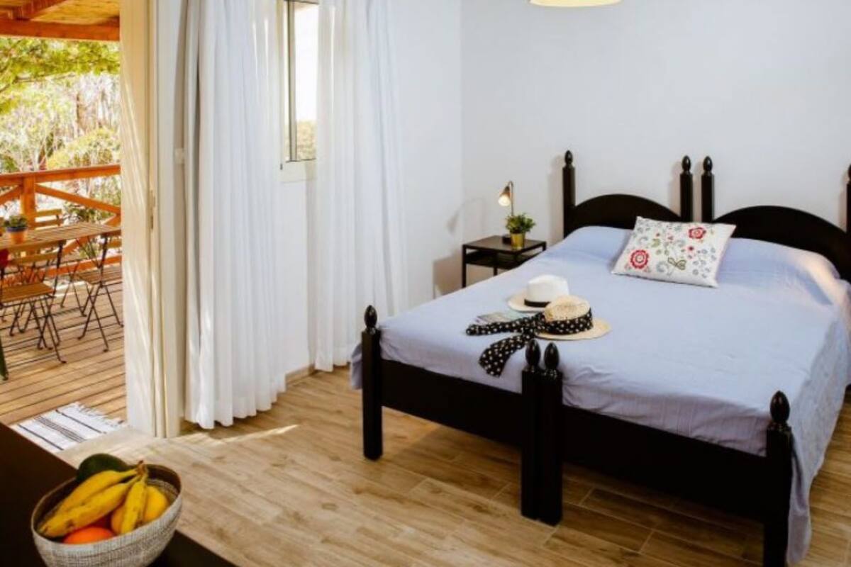 A bedroom with a bed, a nightstand, a window with curtains, a wooden floor, and a view of an outdoor deck.