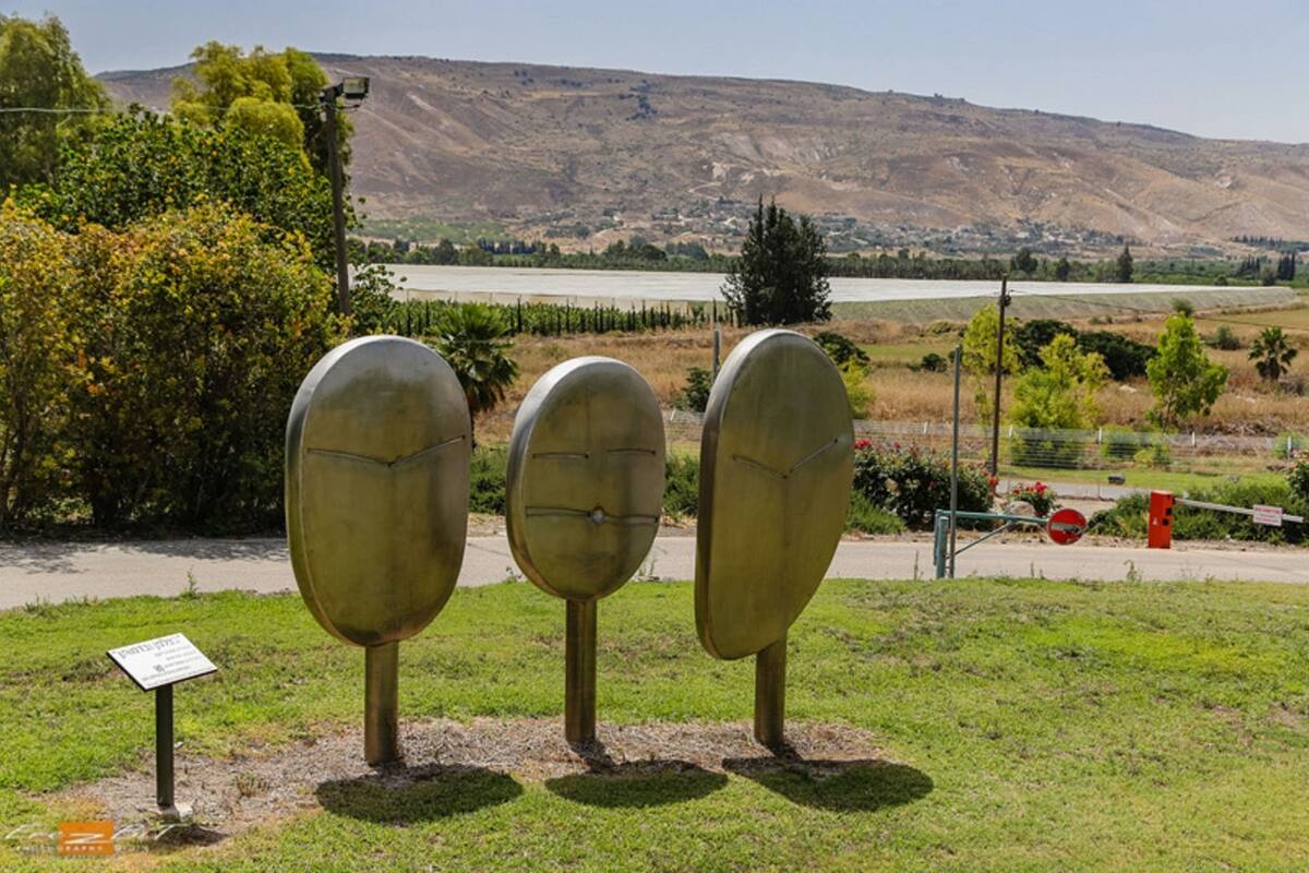 Three metallic sculptures in a grassy area with a backdrop of hills and a body of water.