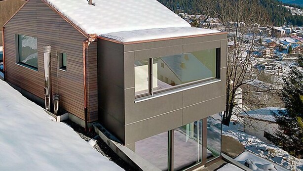 Una casa moderna con techo inclinado, ventanas amplias y vista a un paisaje nevado y un pueblo.