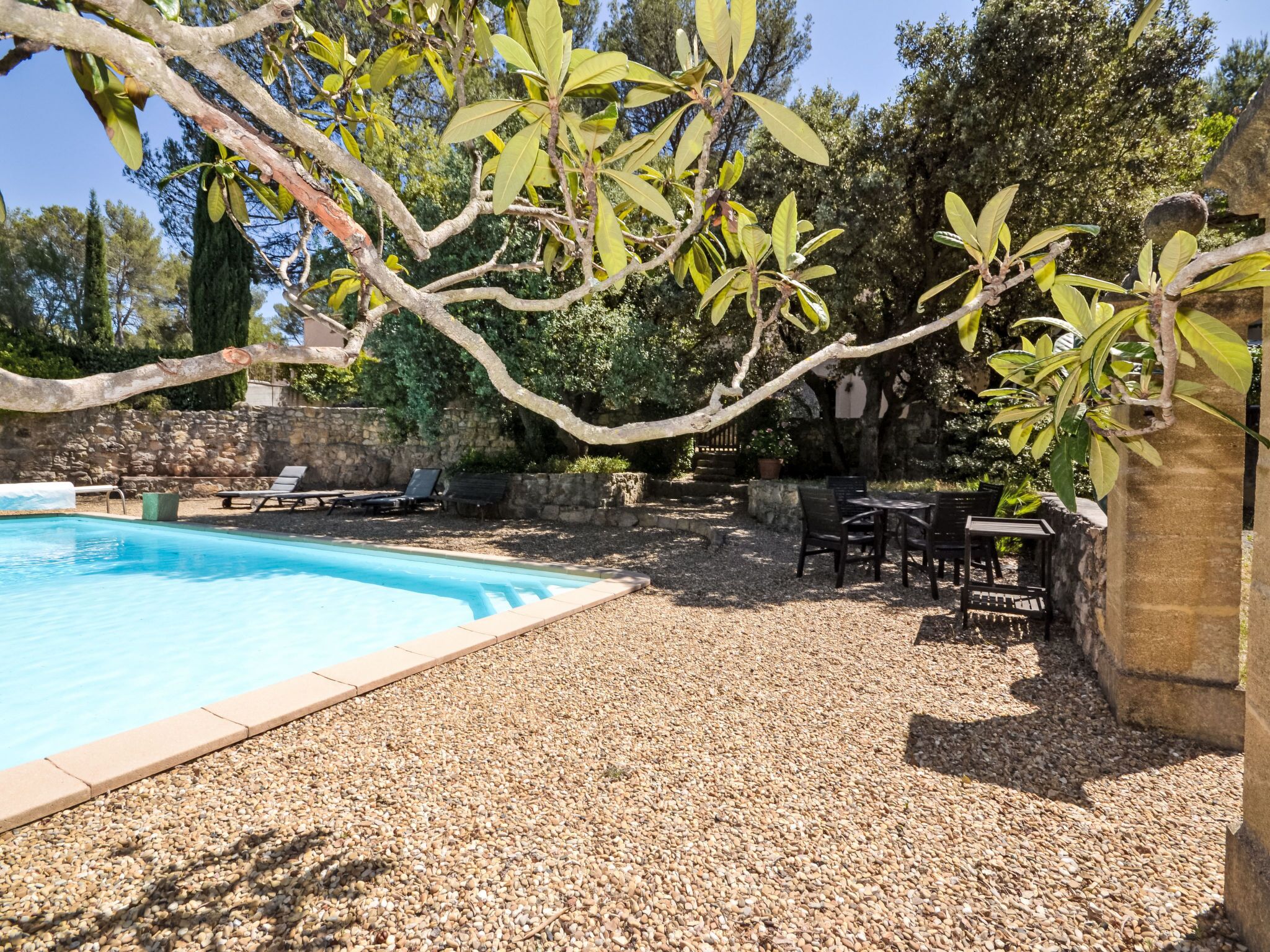 A swimming pool surrounded by a pebble-covered patio with lounge chairs and a stone wall.