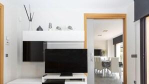 Un soggiorno moderno con una TV a schermo piatto montata su un mobile bianco, una zona pranzo con tavolo e sedie, e vista all'esterno tramite una porta scorrevole.