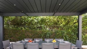 Area pranzo all'aperto con tavolo apparecchiato per sei, circondato da sedie, sotto una pergola con recinzione in metallo e vegetazione sullo sfondo.