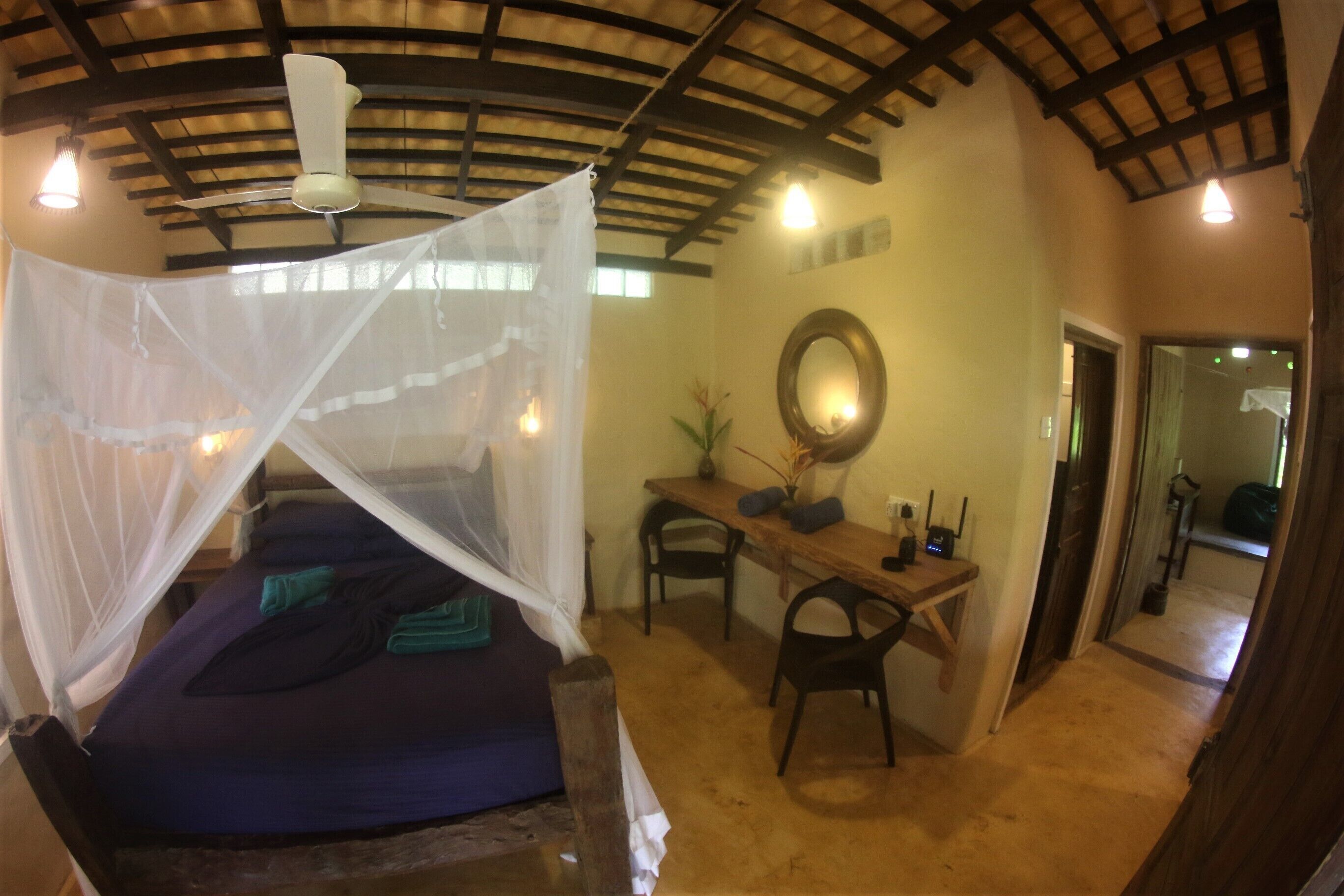 A room with a bed under a mosquito net, a desk with a chair, a mirror, and a small table with a vase.