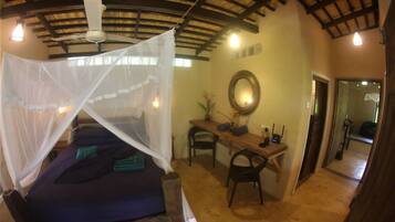 A room with a bed under a mosquito net, a desk with a chair, a mirror, and a small table with a vase.