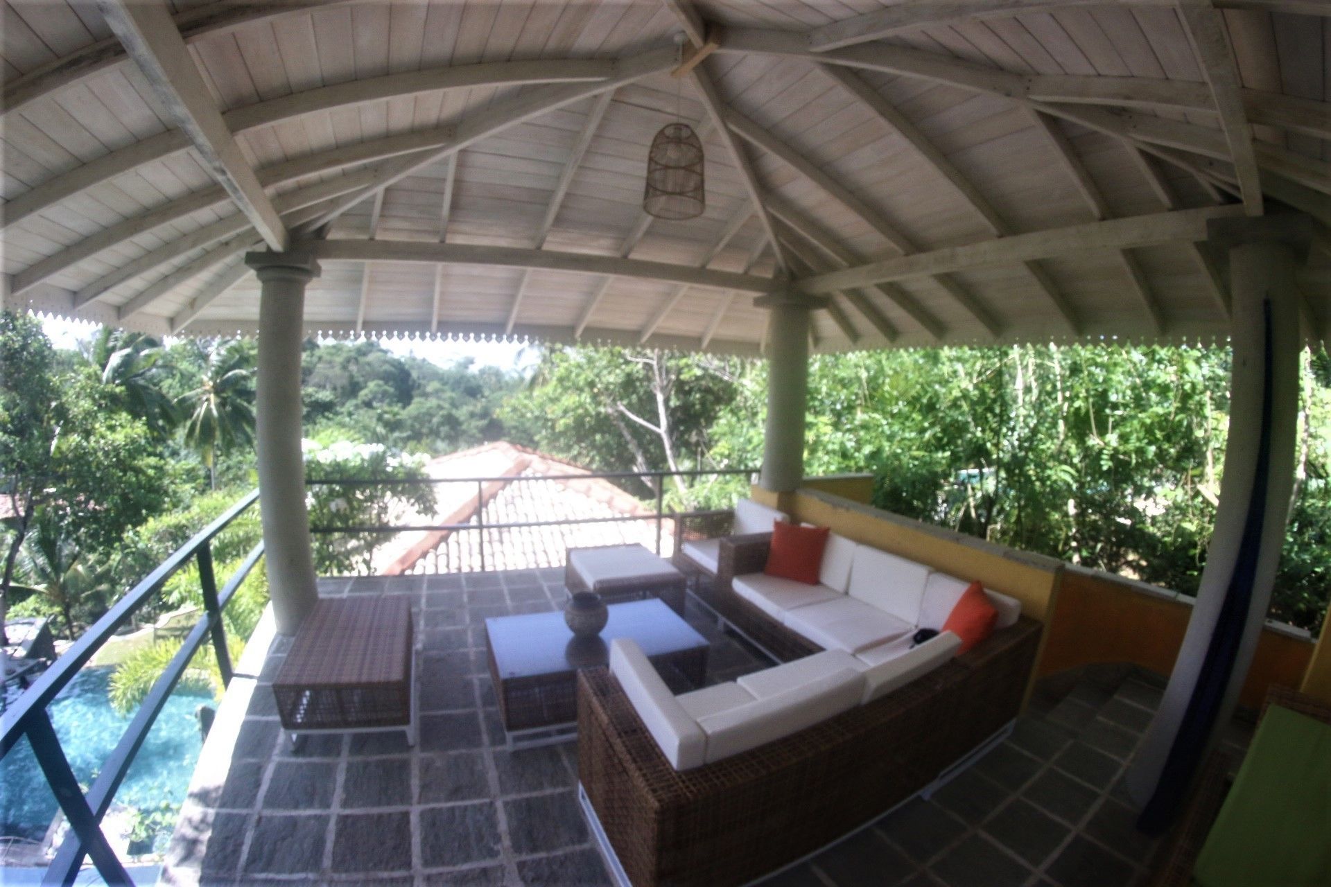 A covered outdoor seating area with a sofa and a table, overlooking a pool and lush greenery.