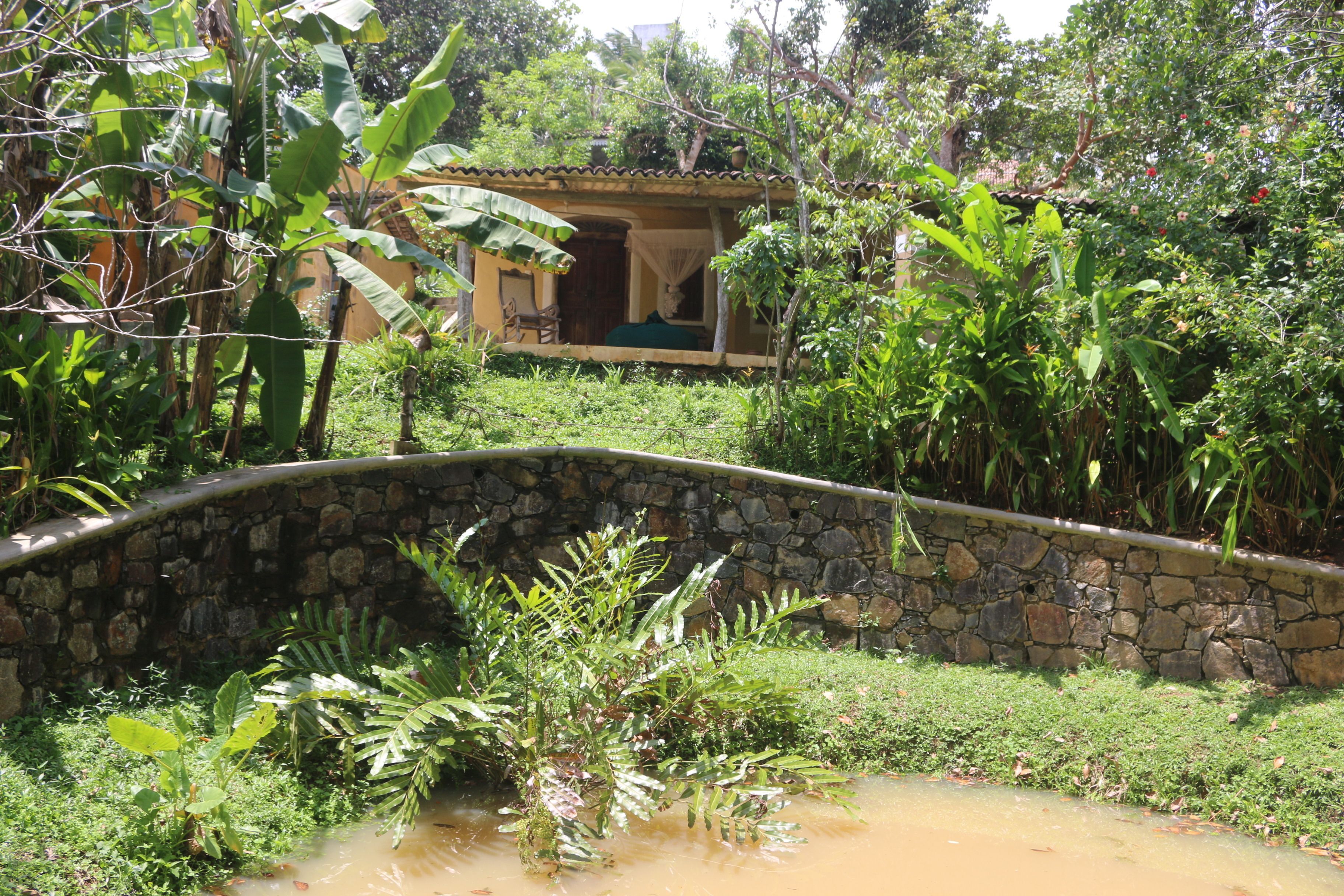A stone bridge over a small stream in a lush garden with a house in the background.