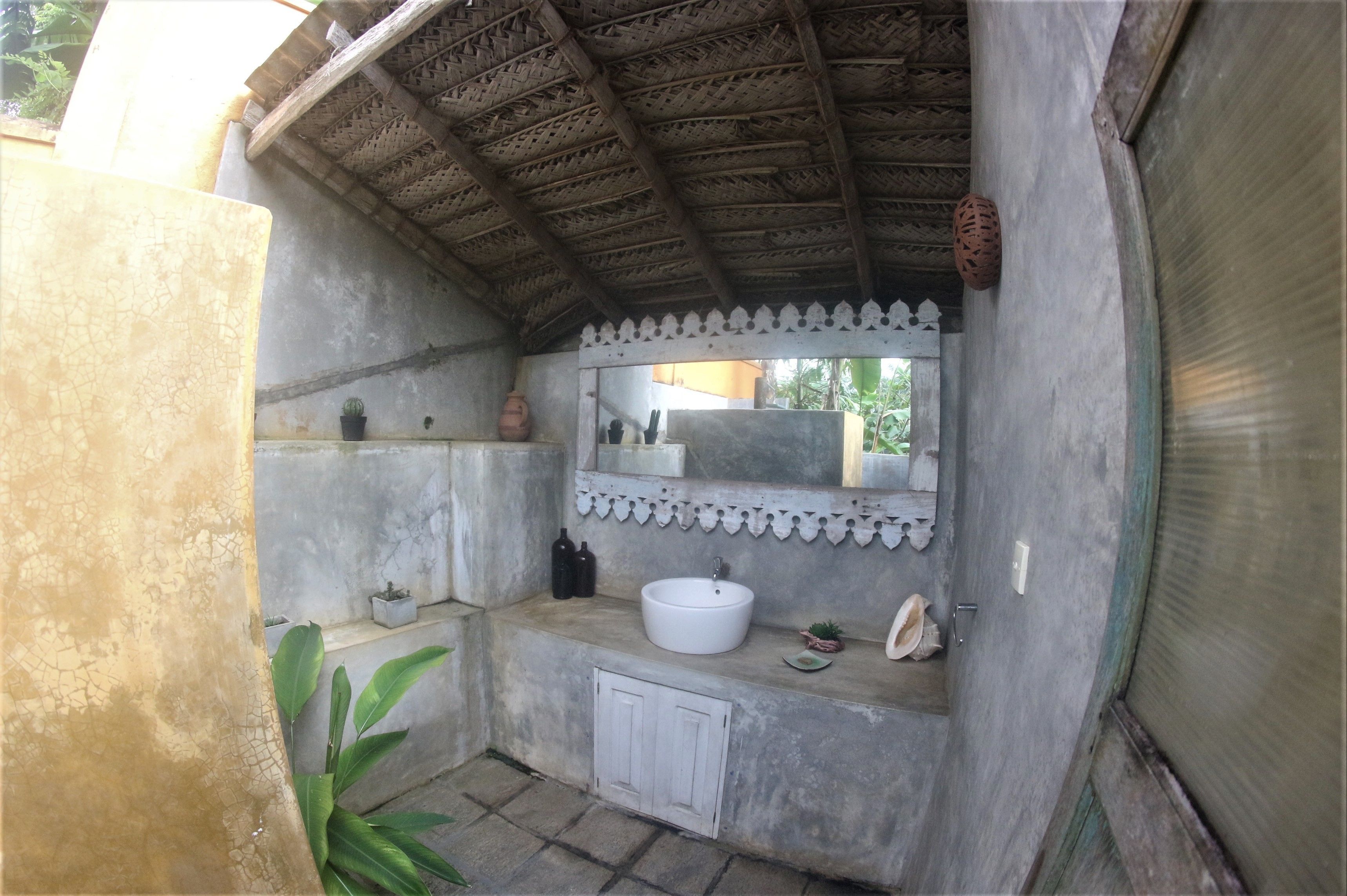 A small, enclosed bathroom with a concrete floor, a white sink, a mirror, and a small shelf with decorative items.