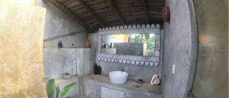 A small, enclosed bathroom with a concrete floor, a white sink, a mirror, and a small shelf with decorative items.