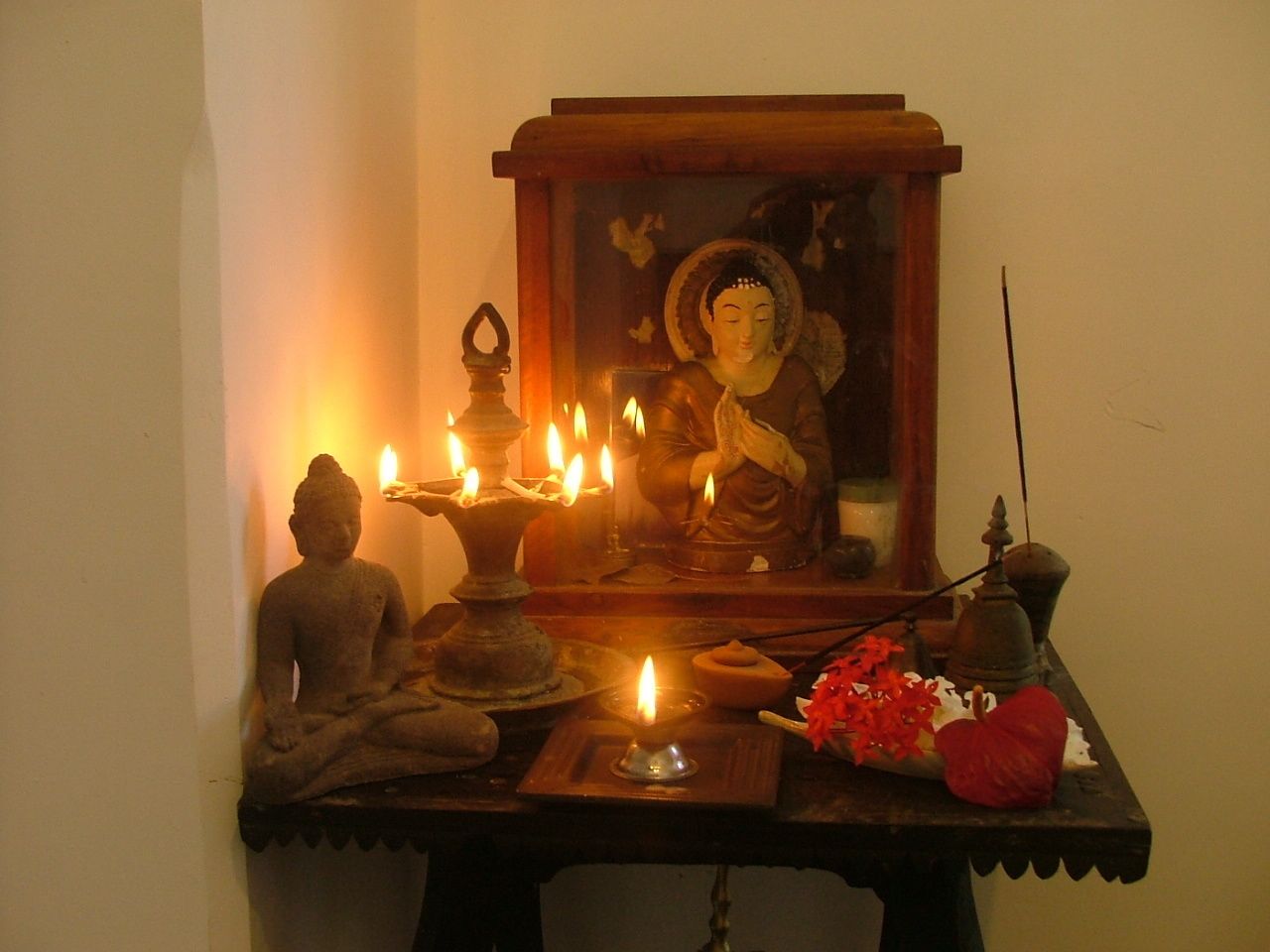 A statue of Buddha with a candle in front of it, a mirror, and a small table with a red flower and a bell.