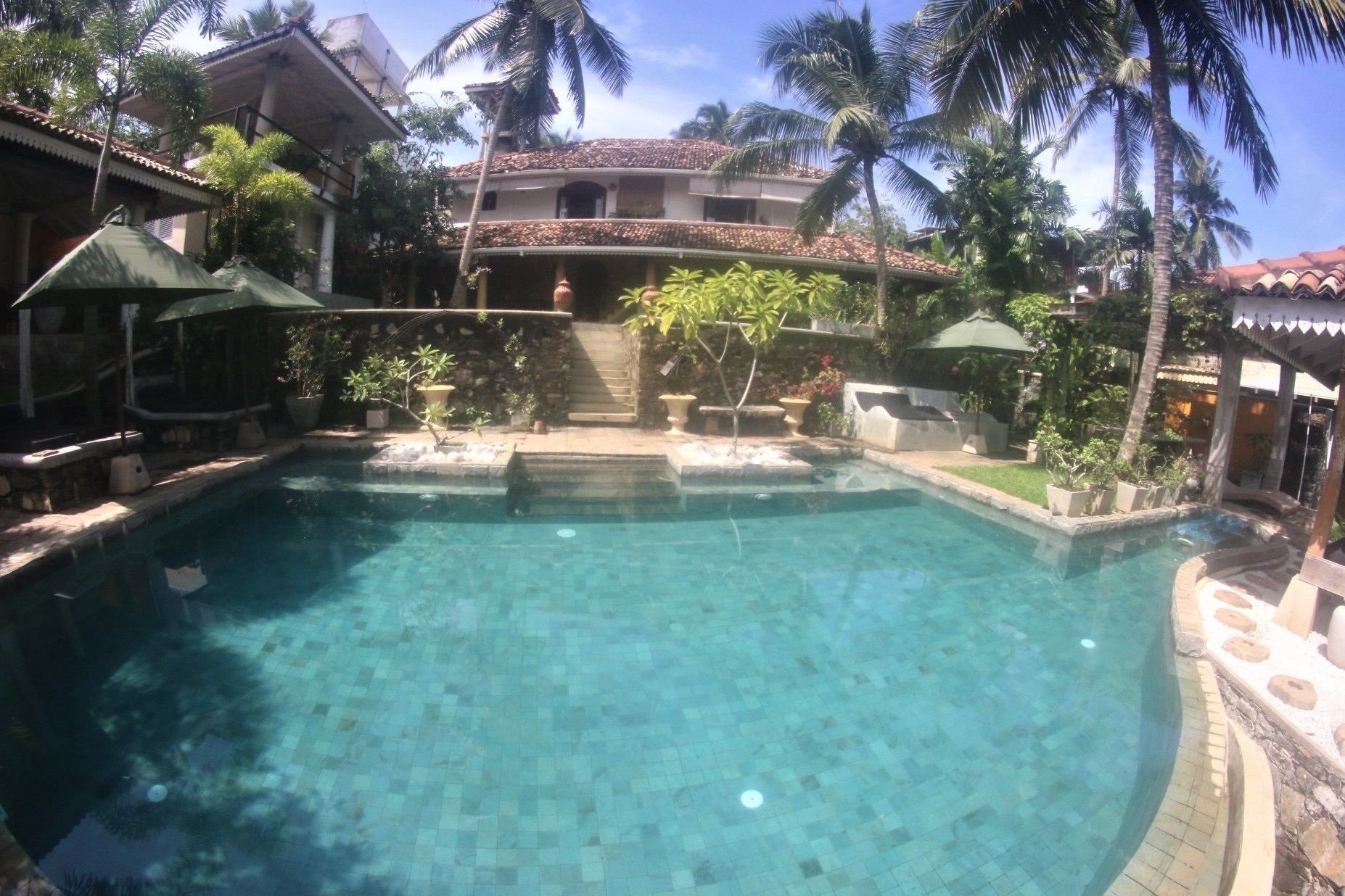 A clear swimming pool surrounded by tropical plants and a multi-story building with a tiled roof.