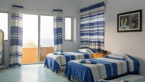 A room with two beds, a small table, and a chair. There is a window with curtains and a view of the outside.