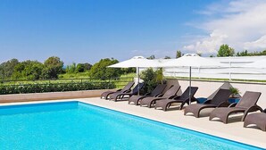 Une piscine entourée de verdure, avec des chaises longues et des parasols, sous un ciel dégagé.