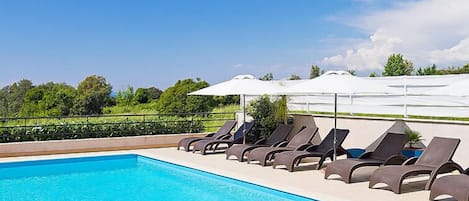 Ein Schwimmbecken mit Liegestühlen und Sonnenschirmen, umgeben von Grünflächen und einem wolkenlosen Himmel.
