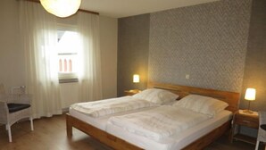 A double bed with white linens, wooden headboard, two bedside tables with lamps, a chair, and a window with curtains. - Haus Irmgard by Interhome (Blankenrath)