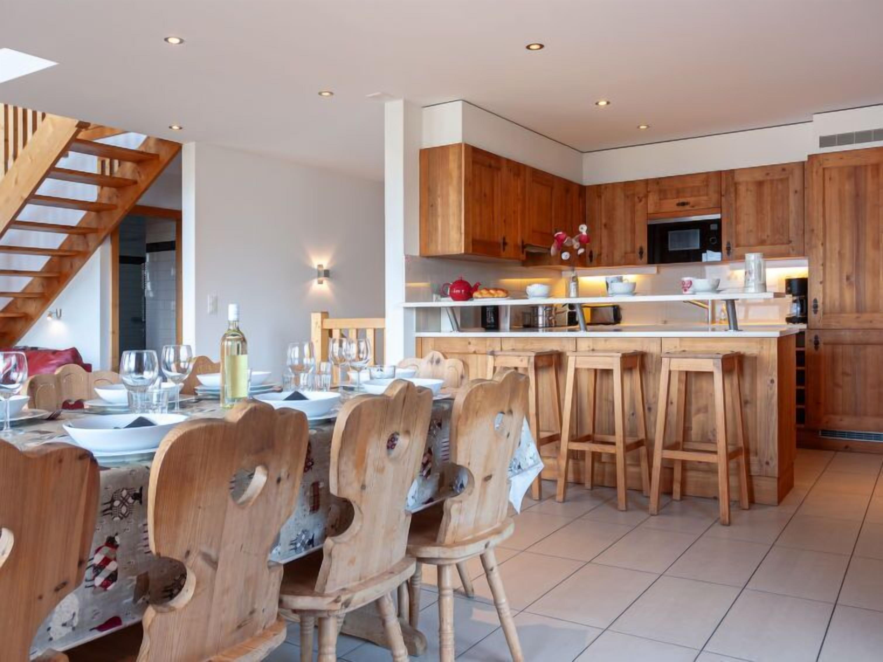 Una cocina amplia con armarios de madera y una isla central, un comedor con sillas de madera, y una escalera que sube a un nivel superior.
