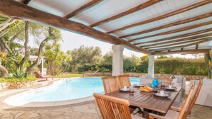 An outdoor pool area with a covered patio, stone flooring, and wooden furniture.