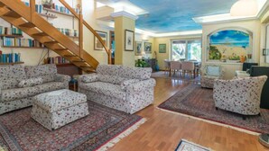 A spacious living room with a wooden staircase, multiple sofas, a dining area, and a large mural on the wall.