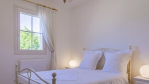 Un dormitorio con cama blanca, una mesita de noche con lámpara, una ventana con cortinas y un radiador.