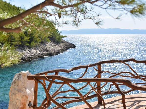 A wooden railing overlooking a clear blue sea with a rocky shoreline and distant mountains.