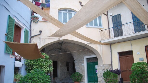 Una calle estrecha flanqueada por edificios, con un arco de piedra, plantas en macetas y un balcón con barandilla de metal.