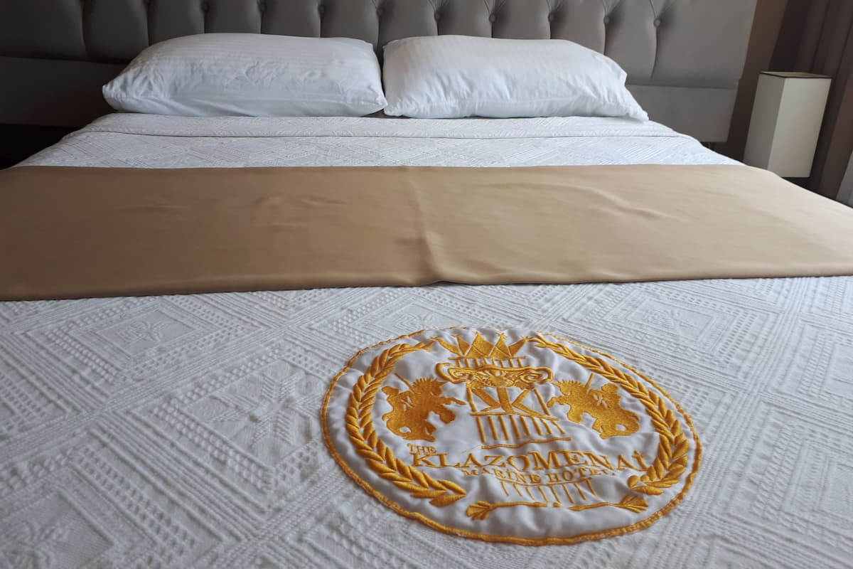 Una cama bien hecha con cubrecama blanco y beige, un emblema bordado en dorado y un cabecero capitonado.