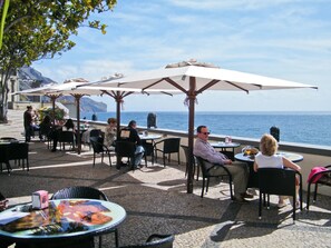 An outdoor café with tables and chairs under umbrellas, overlooking the sea. - OLD TOWN Apartment With Interior Terrace - Barreirinha (Funchal)