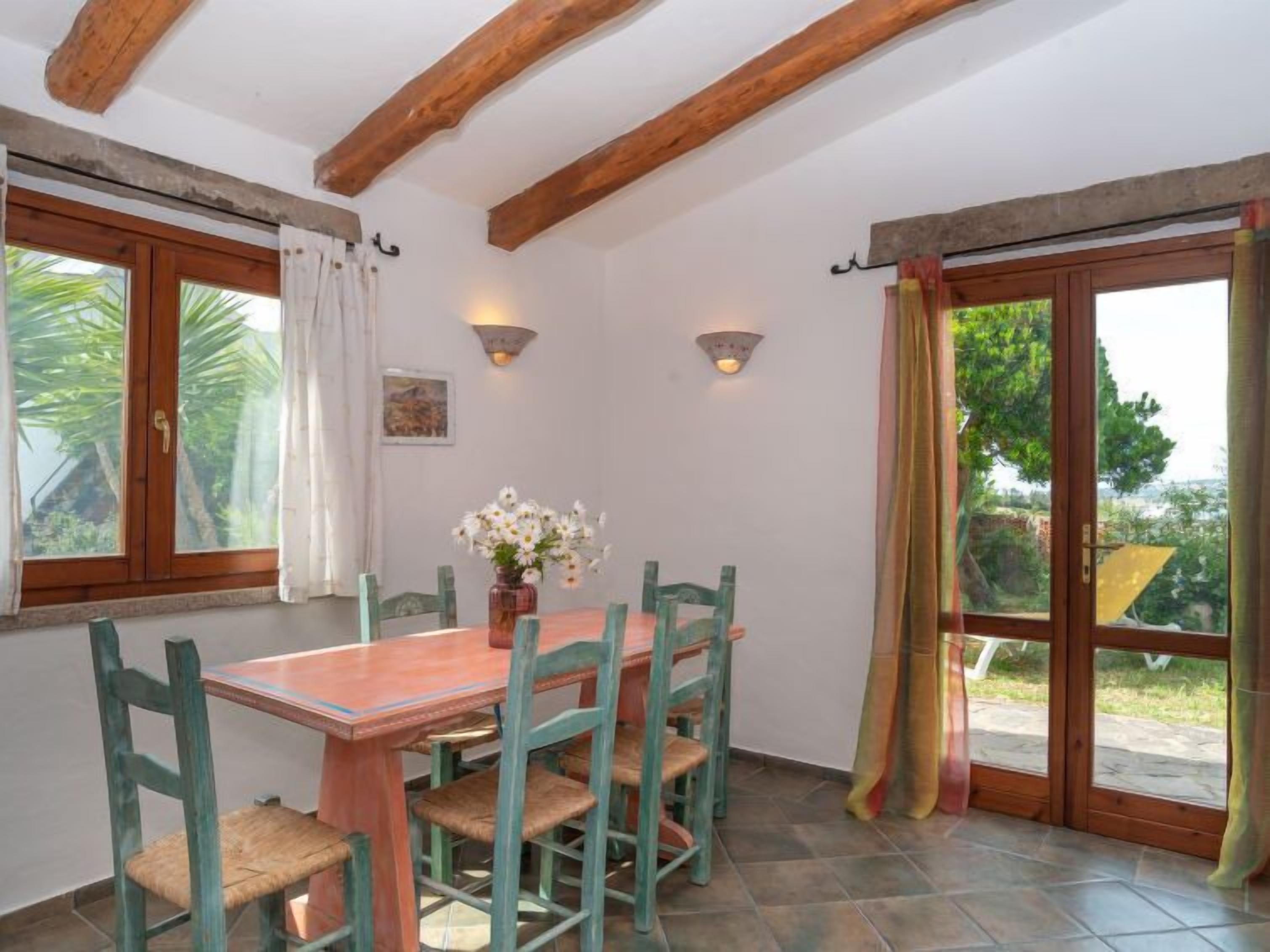 Uma área de refeições com uma mesa e cadeiras de madeira, um vaso de flores e vista para o verde lá fora.