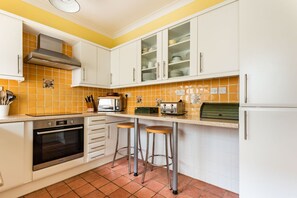 A kitchen with white cabinets, a built-in oven, a toaster, and a bar stool. - Captains Cottage - Four Bedroom Cottage, Sleeps 7 (St Mawes)