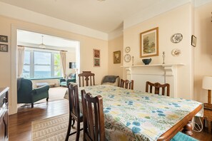 A room with a large dining table, chairs, a fireplace, and a view through a window. - Captains Cottage - Four Bedroom Cottage, Sleeps 7 (St Mawes)