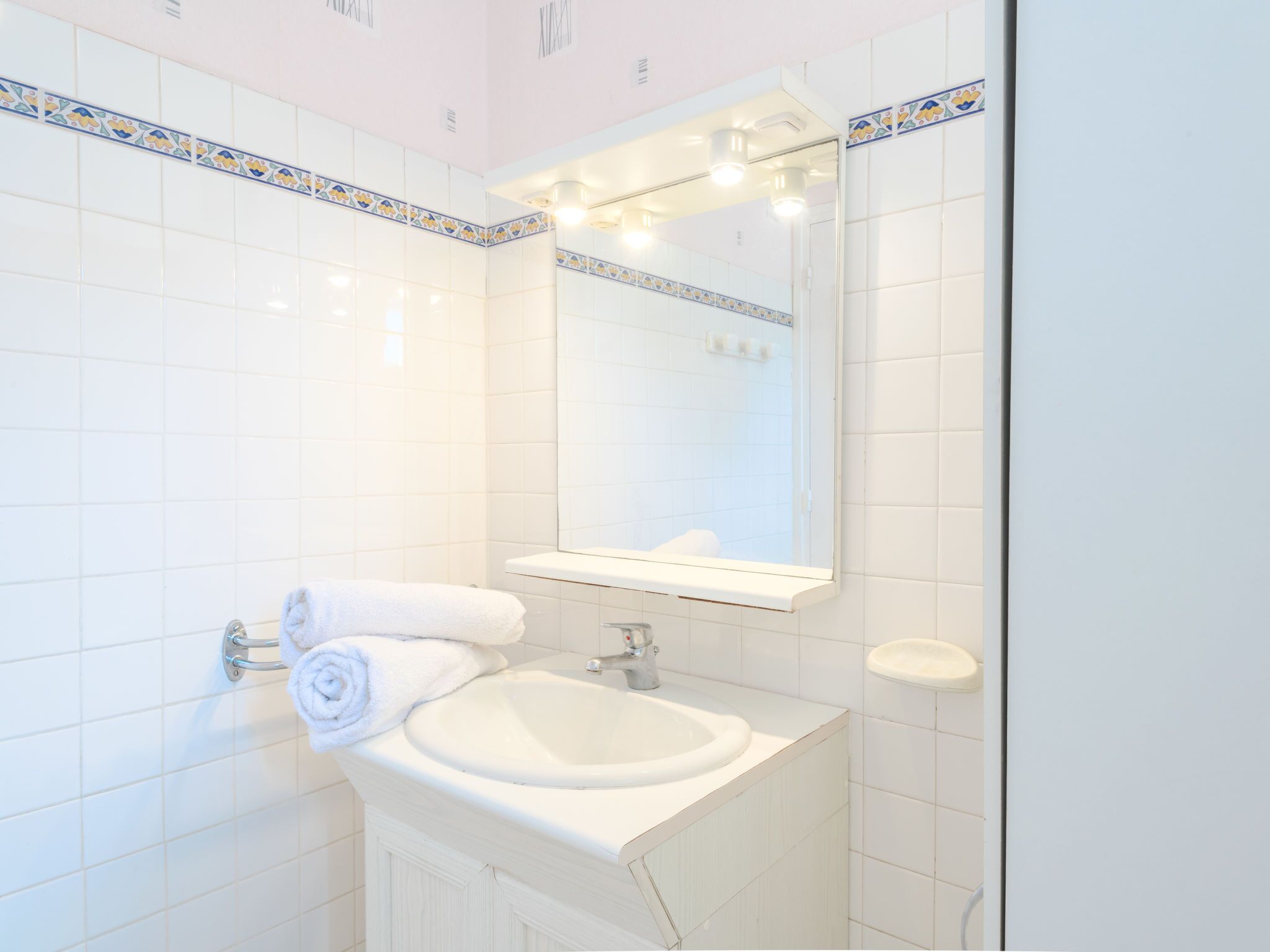 A bathroom with white tiles, a sink, a mirror with lights, and a towel rack.
