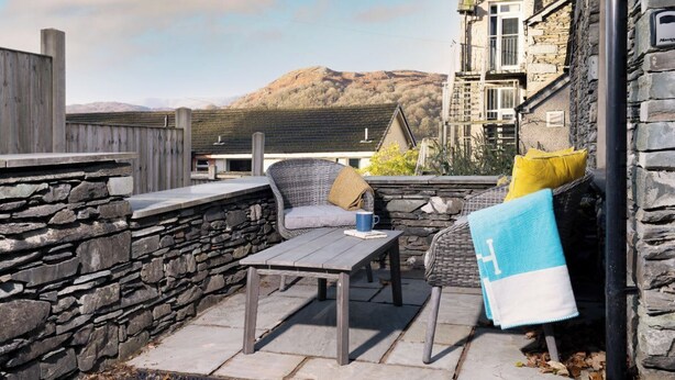 Un espace extérieur aménagé avec des murs en pierre, comprenant une table et des chaises, une serviette suspendue à un crochet, et offrant une vue sur un bâtiment en pierre et des collines en arrière-plan.
