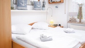 Una cama bien tendida con sábanas blancas y dos toallas dobladas, una lámpara de noche y una mesita con una lámpara y un libro.