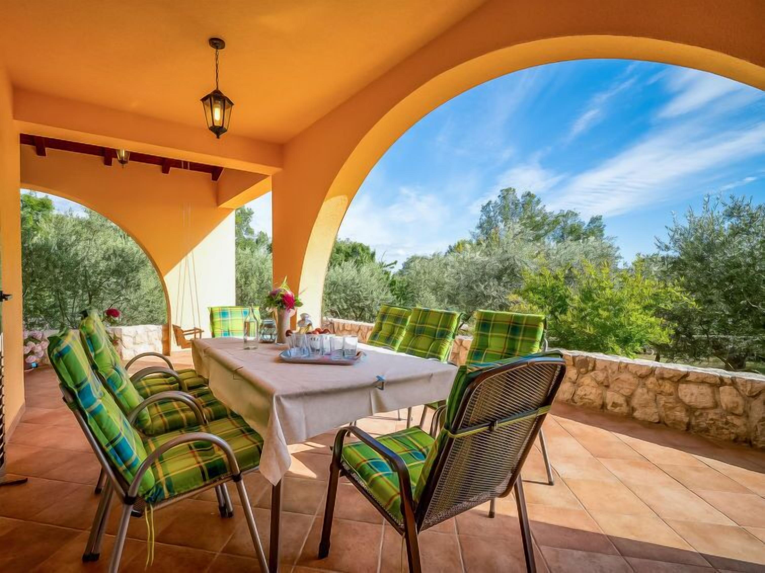 Un patio exterior con una mesa dispuesta para cuatro personas, rodeado de sillas con cojines a cuadros, bajo un gran arco.