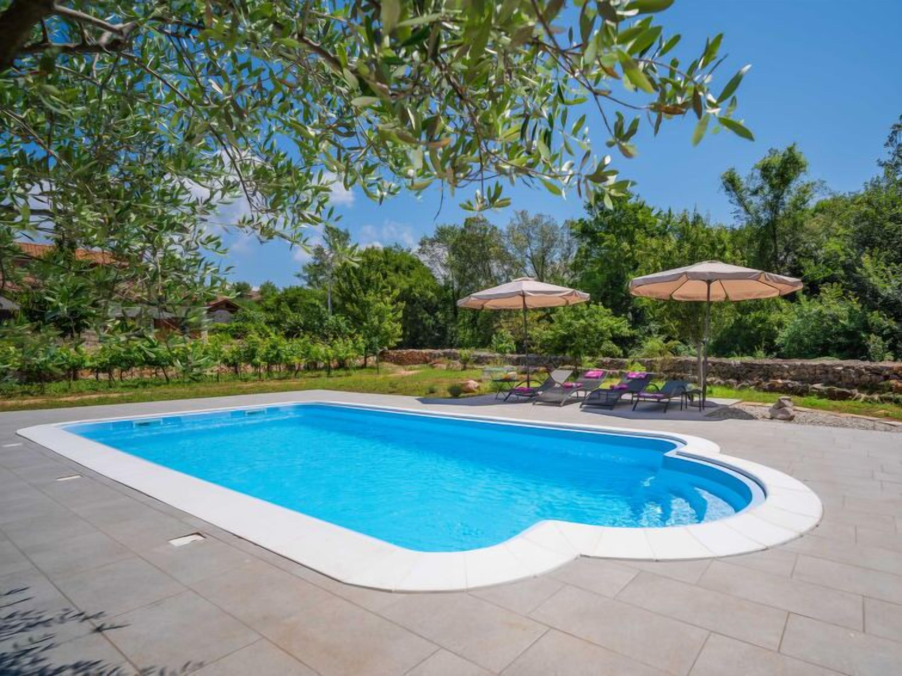 Une piscine rectangulaire bordée de blanc, entourée d’une terrasse dallée. On aperçoit deux parasols et des chaises longues à l’arrière-plan.