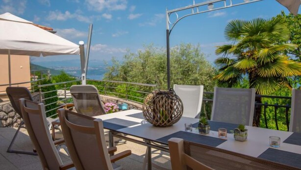 Eine Terrasse mit Tisch und Stühlen, einer Ziervase und Blick auf das Meer und viel Grün.
