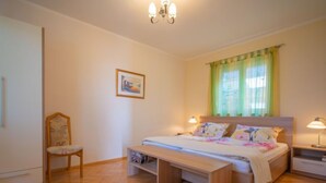 Ein Schlafzimmer mit einem Bett, Nachttischen, einem Sessel, einer Bank, einem Bild an der Wand und einem Fenster mit Vorhängen.