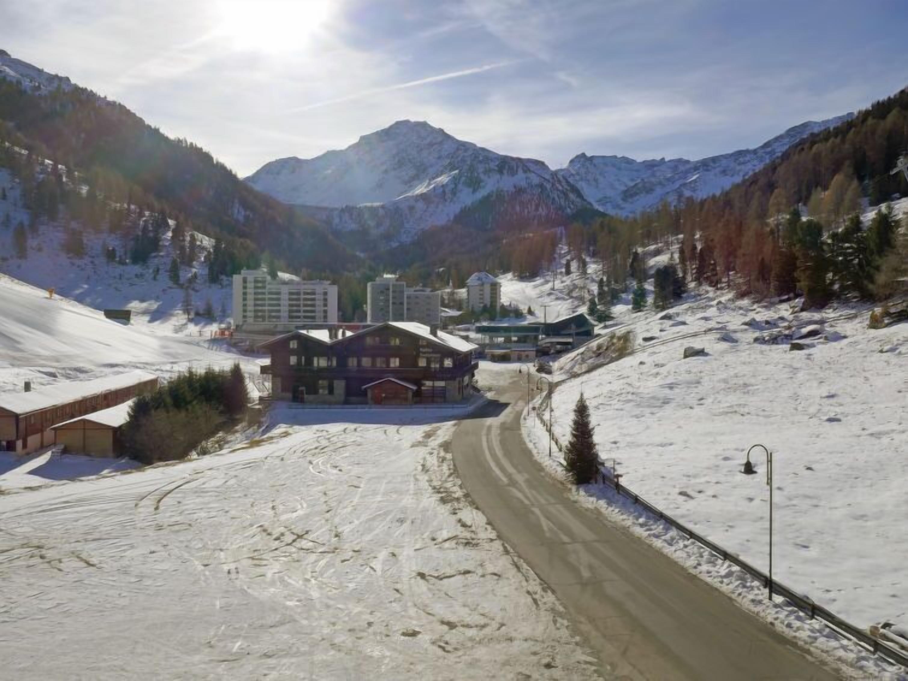 A snow-covered mountain landscape with a building in the foreground, a road, and a few other structures in the background.