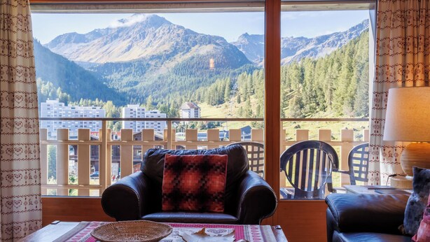 A balcony with a leather sofa and armchair, a table with a woven basket and decorative items, and a view of mountains and buildings. - Dents Rousses B4 by Interhome (Siviez-Nendaz)