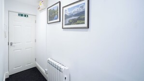 Un couloir avec une porte blanche, deux tableaux représentant des paysages, un panneau indiquant la sortie de secours et un radiateur.